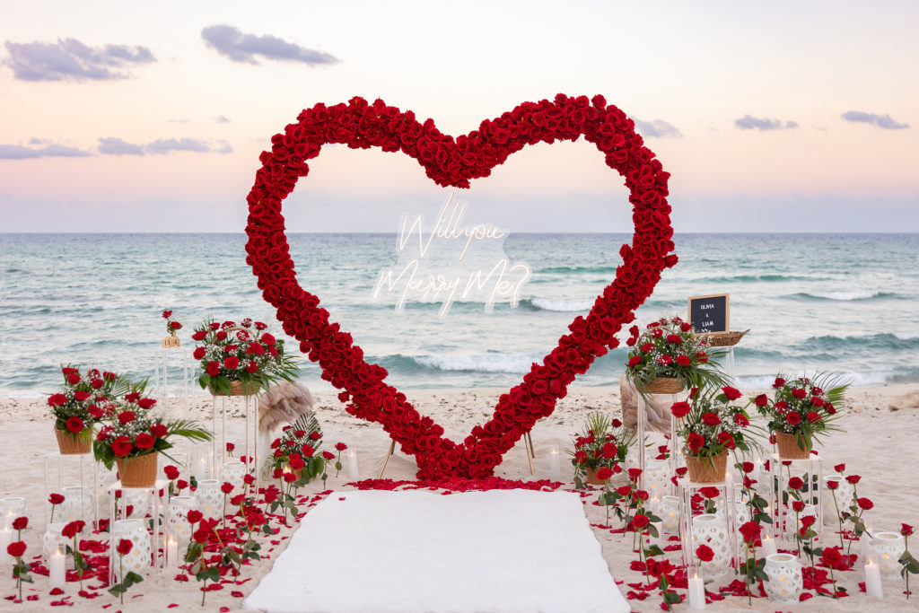 A red heart-shaped floral arch on a beach at sunset with roses and candles lining the aisle.