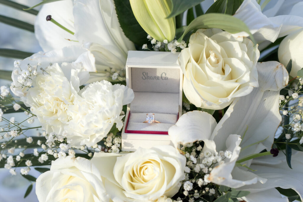 Engagement ring in an open box surrounded by white roses and lilies.