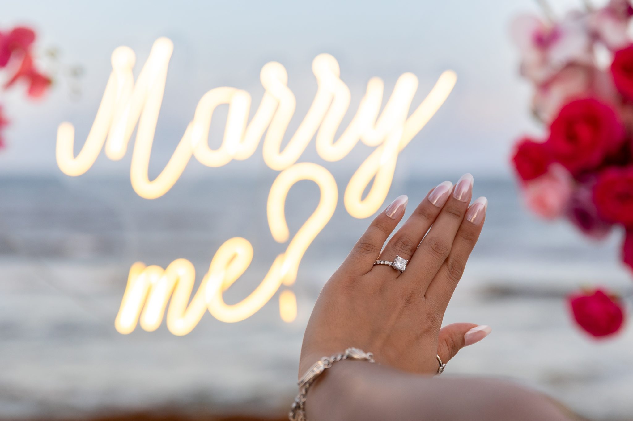Engagement ring in front of a glowing Marry Me sign on the beach