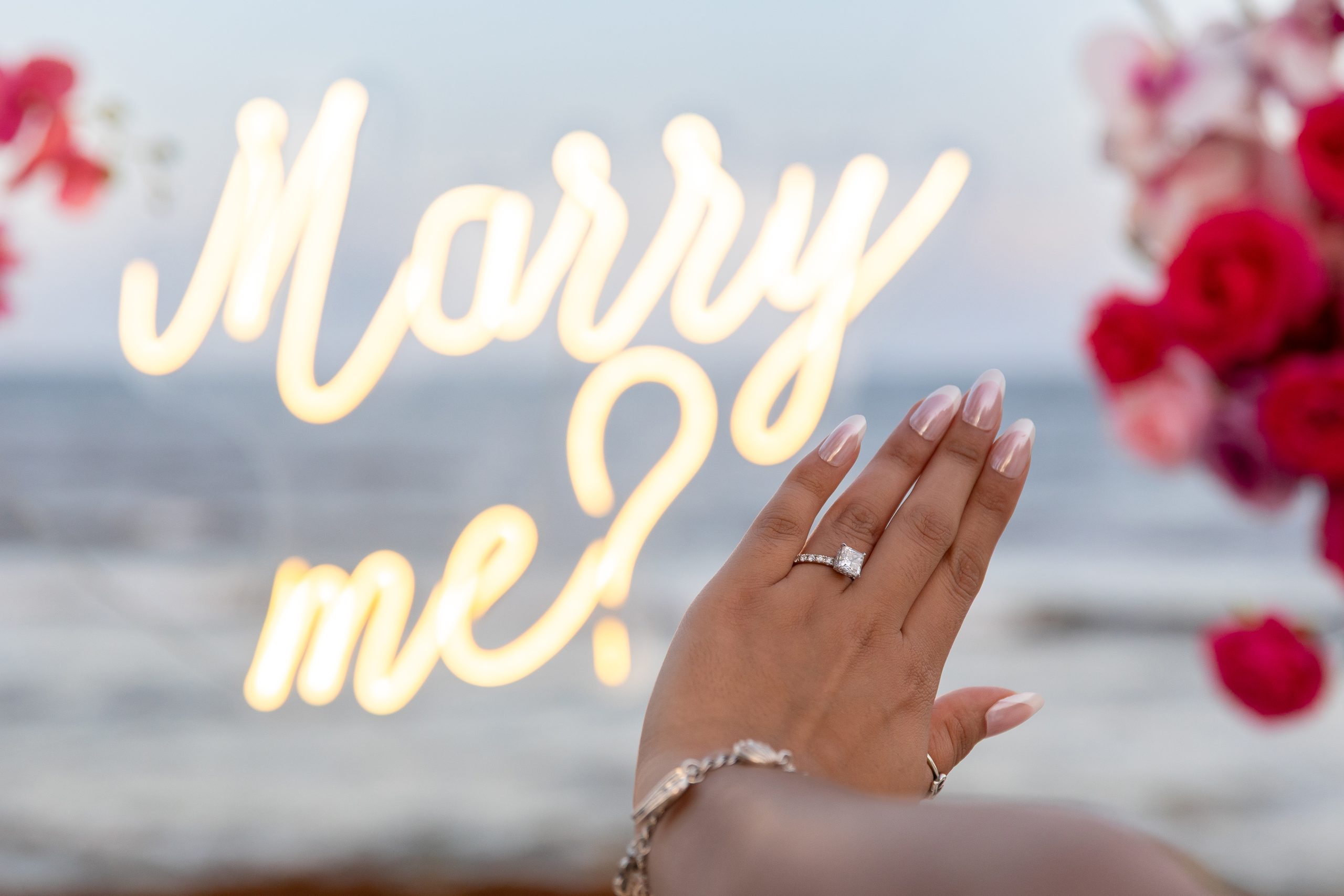 Engagement ring close-up in front of a glowing “Marry Me?” neon sign on the beach