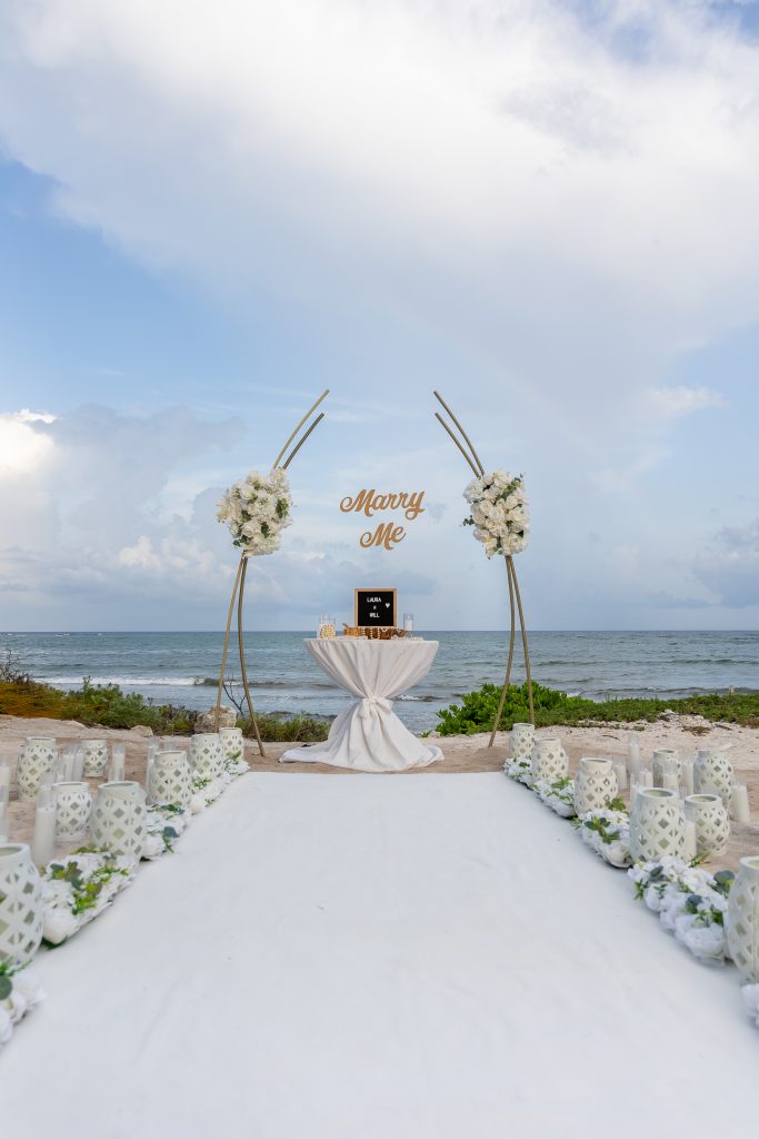 White floral proposal arch with "Marry Me" sign, candles, and lanterns overlooking the ocean