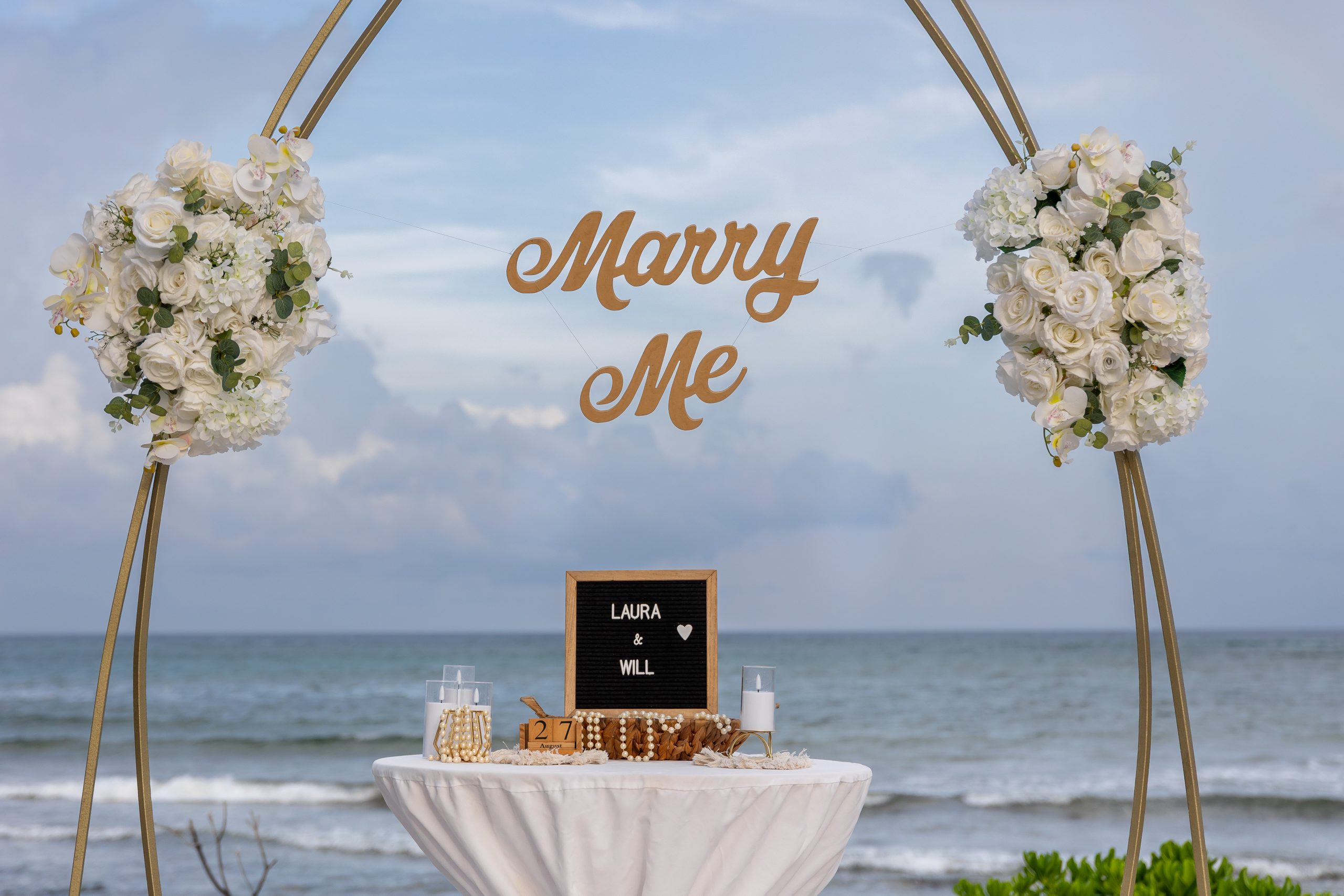 Close-up of “Marry Me” sign with white rose arrangements and personalized message by the beach