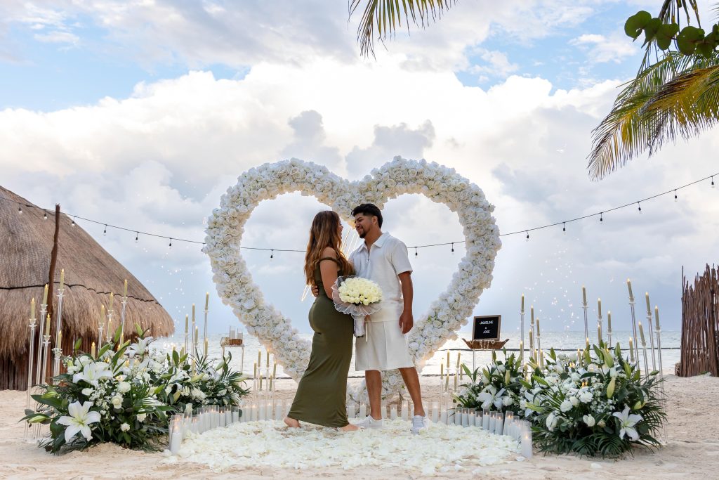 Elegant beach proposal setup with oversized heart arch made of fresh white roses, candles, and floral arrangements at Acamaya Weddings Mexico