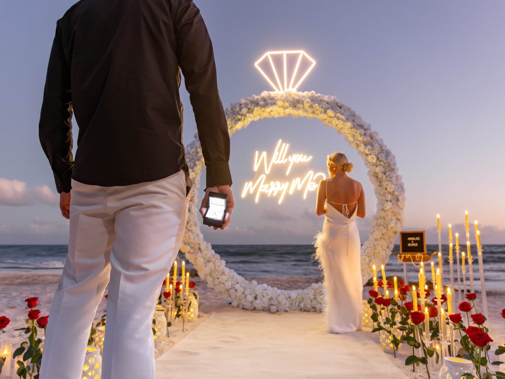 Man holding engagement ring walking toward partner at a beach proposal setup in Playa del Carmen.