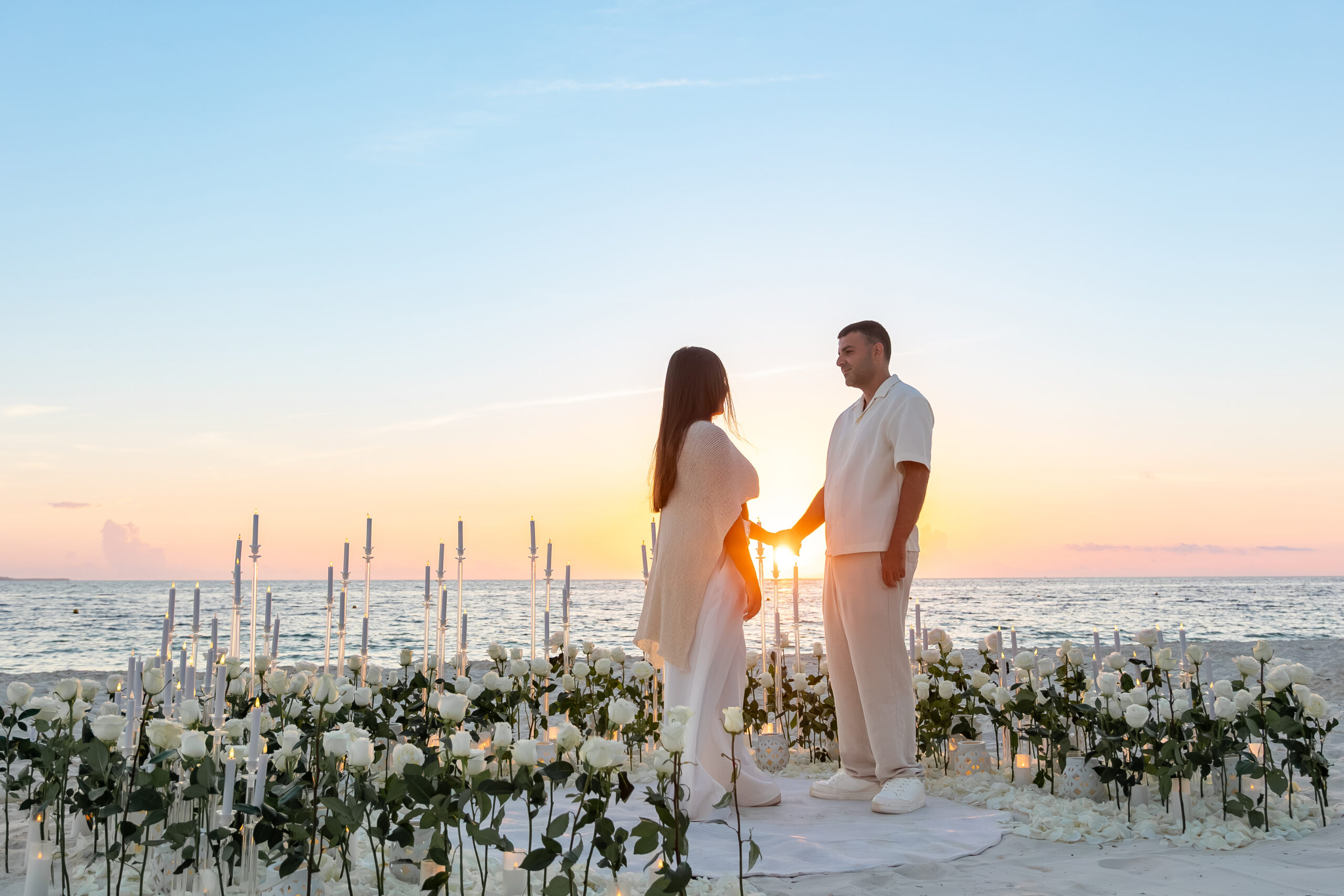 Couple holding hands during a sunrise beach proposal at RIU Caribe in Cancun, surrounded by fresh white roses