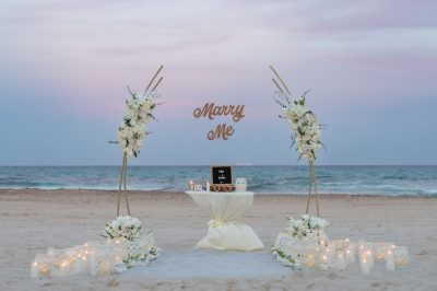 Intimate beach proposal setup with white floral arch, candles, and Caribbean ocean view in Riviera Maya